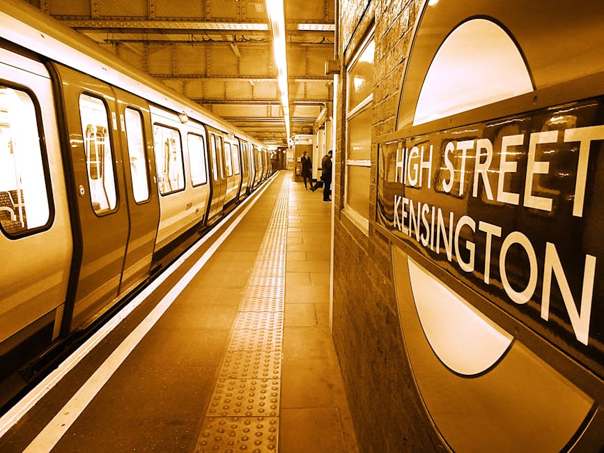 Indoor view of an underground train station platform at High Street Kensington, showing clean, polished ceramic tile flooring with tactile paving strips near the edge, illuminated by overhead fluorescent lighting. The platform features a modern train with closed doors on the left, and on the right, a wall mounted sign displaying 'High Street Kensington' within a circular station emblem. The wall behind the sign is made of brick or stone, and a few passengers are visible in the distance. This setting exemplifies well-maintained surface cleaning and hygiene, as provided by Kennington Carpet Cleaning, highlighting their expertise in deep cleaning and sanitisation for residential and commercial spaces.