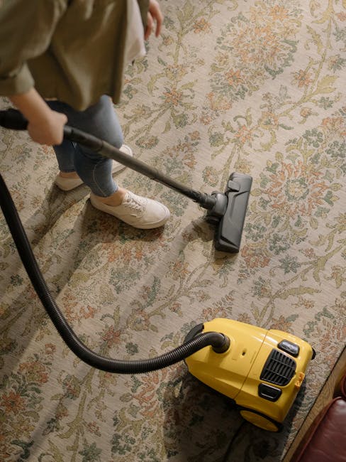 A person using a handheld vacuum cleaner to deep clean a traditional patterned area rug with floral motifs in a domestic setting. The rug, placed on a hard floor surface, displays detailed designs in shades of beige, green, and orange. The individual is wearing casual clothing, including white sneakers and beige pants, and is actively vacuuming the rug to remove dust and debris, demonstrating surface cleaning and maintenance typical of professional residential cleaning services by Kennington Carpet Cleaning. The scene is illuminated with soft indoor lighting, emphasizing the clean and well-maintained appearance of the rug during the cleaning process.