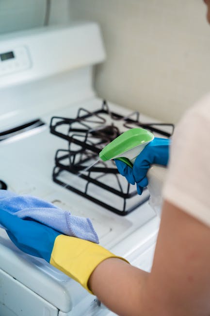 A person wearing blue and yellow cleaning gloves is holding a spray bottle and a cloth, preparing to clean a white stovetop with black metal burners in a kitchen. The stovetop and surrounding beige wall are visible, with the surface appearing clean and dry. The scene is well-lit with natural or artificial light, and the focus is on the surface cleaning process. The image illustrates domestic cleaning and surface sanitisation, consistent with the services of Kennington Carpet Cleaning as referenced in the context of the Kennington Park deep cleaning guide for households.